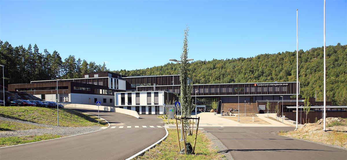 Stor trekledd bygning med asfalt foran og skog bak. Foto. - Klikk for stort bilde